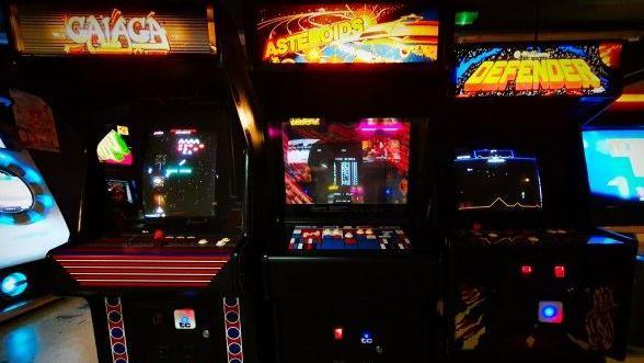 Three arcade machines with lit-up signs on the front and the screens are on. The room is dark and the lights are bright.