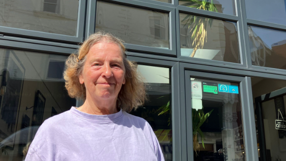 Sandy Peters, smiling at the camera, in front of the glass doors to the Sopranos restaurant wearing a purple t-shirt. 