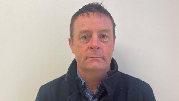 A man, Raymond Gormley. Pictured from the shoulders up, he is wearing a blue shirt and a navy jacket. He has brunette hair.