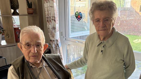 Gordon Cilgram with white hair and wearing glasses a dark green waistcoat and beige shirt sits by the side of his wife Joan who has white hair and glasses and green top standing beside him 