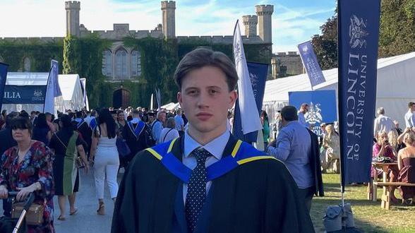 James Harrison at his graduation ceremony stood outside. He's wearing a black cape with blue and yellow accents. He is in front of a bustling crowd of other graduates.