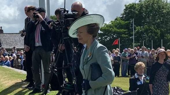 Princess Anne dressed in pale green coat and hat at the Tynwald Day ceremony. Press representatives with cameras are standing behind her with crowds in the background.