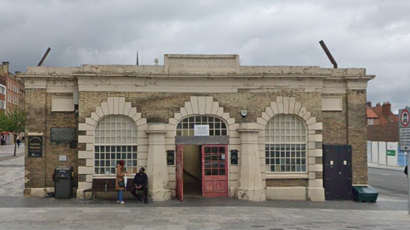 An outside view of the Stockton Shambles building. It has open red doors in the centre of stone archways with windows in them. Two people are near the bench to the left of the front doors.