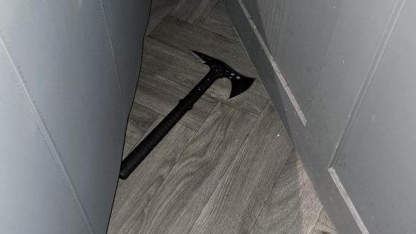 A double-headed axe, with a black handle. It is on a wooden floor between a grey wall and the grey back of what appears to be a sofa.