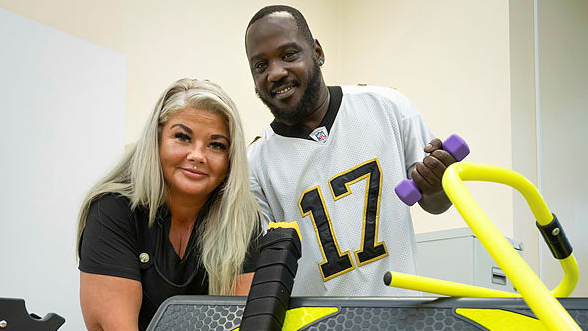 Anita and Matthew are pictured with the equipment. Anita has long blonde hair and a black top on. Matthew has an American football top on and they are both standing behind some of the new equipment.