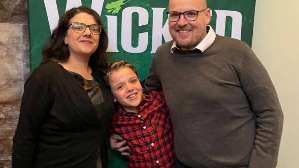 Three people stand in front of a poster of the play Wicked. A woman in a dark cardigan and black hair, a boy in a red and black checked shirt and a man with a bald head and black spectacles wearing a brown jumper.