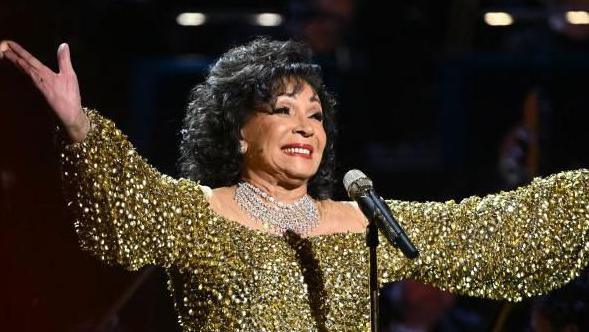 Gold sparkly dressed Dame Shirley Bassey grinning after a performance, she has curled black hair and has her hands outstretched 