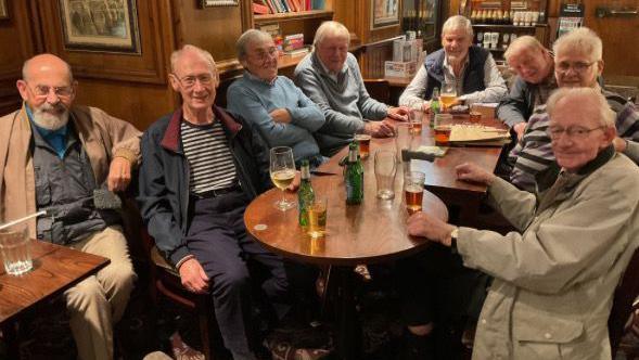 Eight elderly men sat around wooden tables with pint glasses and green beer bottles