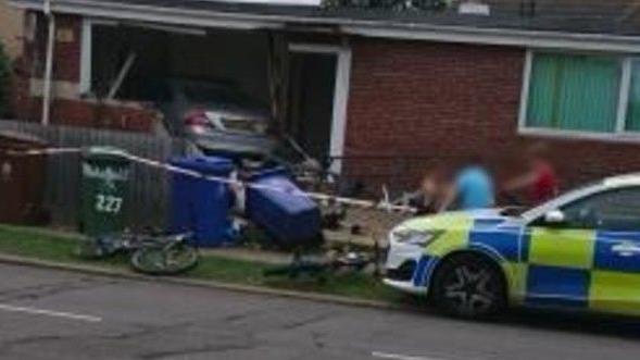 A view of a badly damaged house with a silver Mercedes car inside it. The wall of a house has been largely destroyed. There is a police car in the foreground, along with a number of wheelie bins, one lying on its side.
