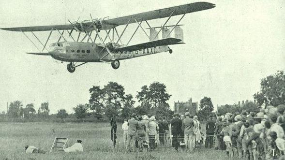 A black-and-white photograph of a Hercules aircraft approaching Brockworth airfield in 1932. The plane is still airborne, descending over a wide grassy field. On the right side of the image, a crowd of spectators stands with their backs to the camera. Two people dressed in white are lying on the grass in the foreground, apparently trying to get a better view. The scene captures the anticipation of the landing.