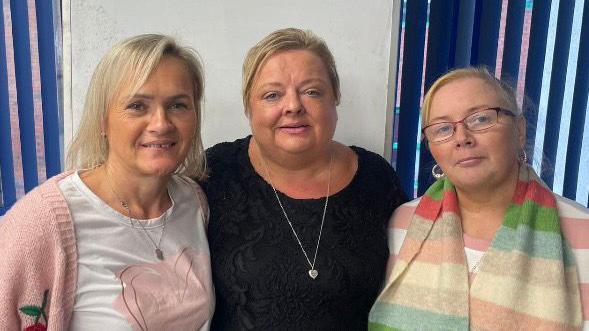 Suzanne O'Hara (l), Linda Harvey (m) and Annette Park (r). Suzanne has shoulder-length blonde hair, wearing a white t-shirt with pink heart, necklace and pink cardigan. Linda has short blonde hair, wearing a long silver heart necklace and black top. Annette has blonde hair tied back, wearing glasses, hoop earrings, multi-coloured scarf and pink and white striped top.