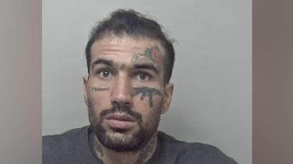 Custody photo of Fayaz Khan, who has short dark hair, a beard and moustache. He has tattoos on his face, including one of an AK-47 gun beneath his left eye. He is wearing a grey jumper.