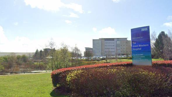 Hospital entrance under sunny sky showing green lawns, pond and large entrance sign.