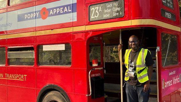 A man in a conductor's uniform and yellow high-viz vest stands on the back door of a traditional routemaster red bus. He is smiling at the camera
