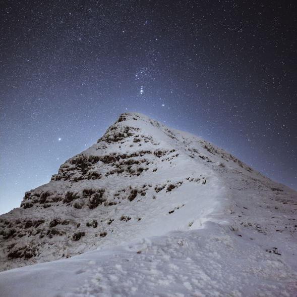 Starry skies above Pen Y Fan seen from north ridge. The mountain is covered in snow and the sky is a very dark blue.