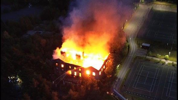 Drone footage of a two-storey 18th Century manor house engulfed in orange flames, while jets of water from fire engines are aimed at its roof.
