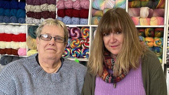 A woman with short hair and a grey jumper and a woman with long hair and a purple jumper stand in front of shelves stocked with colourful balls of yarn.