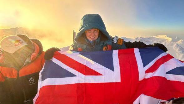 Adriana Brownlee stands and looks down at her Union Jack flag while on the peak of a mountain in a blue snowsuit and yellow hat