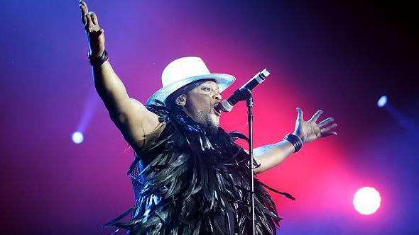 D'Angelo, wearing a white hat and a boa with black feathers around his neck, performs on a stage with bright pink lighting. He is raising his arms in the air.