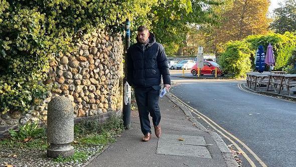 Stuart Worby arriving at court wearing a black puffa jacket. He has his head turned away from the camera, and is wearing a suit, with brown shoes. He is walking up a road, and a car park can be seen in the background along with some picnic tables to the left. 