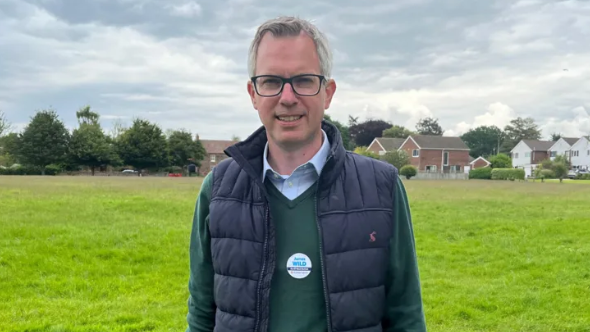 James Wild stood in a field with houses in the background. He is wearing a light blue shirt, green jumper and an open black gilet. He has short grey hair and back glasses and is smiling. 