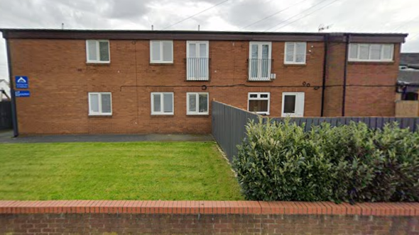 Westmead carehome in Castleford sits behind a wall and lawn. A fence runs through the grass which features a hedge in front of it.