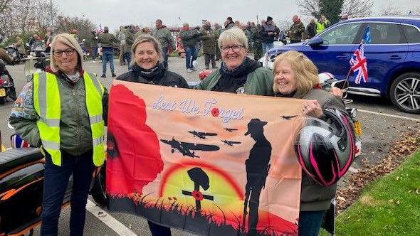 Four women in parkas hold an orange flag reading 'Lest We Forget' with poppies and soldiers