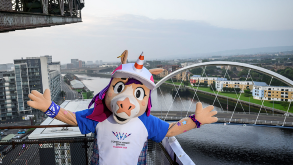 A anthropomorphic unicorn, with a traffic cone for a horn and wearing blue and white Scotland top for the Commonwealth Games. The mascot is posing with a view of Glasgow and the river Clyde stretching behind them.