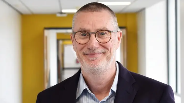 A man with short grey hair and a beard and glasses. He is wearing a striped shirt and dark jacket