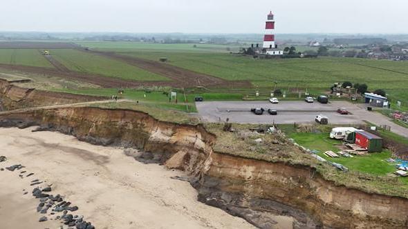 Drone aerial shot of the coast line in Happisburgh. You can see some buildings, a car park and a red and white striped lighthouse.