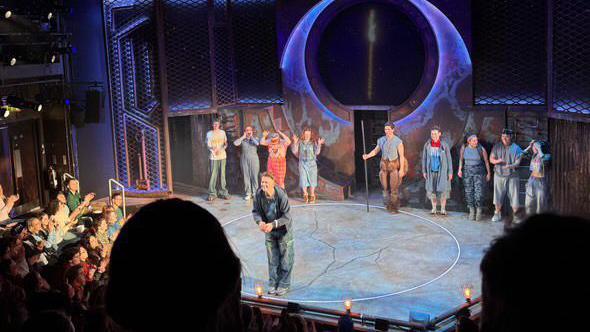 A man bowing on stage whilst other performers stand in a line behind him. People are applauding him in the crowd.