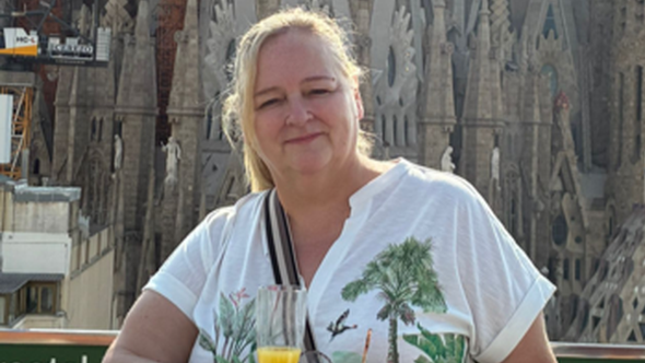 Alison has blonde hair in a ponytail, wears a white t-shirt with a palm tree design. She has a crossbody handbag on and holds a champagne flute with what appears to be orange juice inside. In the background is a large ornamental building which appears to be a cathedral.