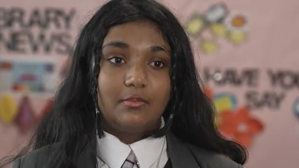Emaan has long black hair with two mini braids in the front. She wears a grey balzer, white shirt and tie.