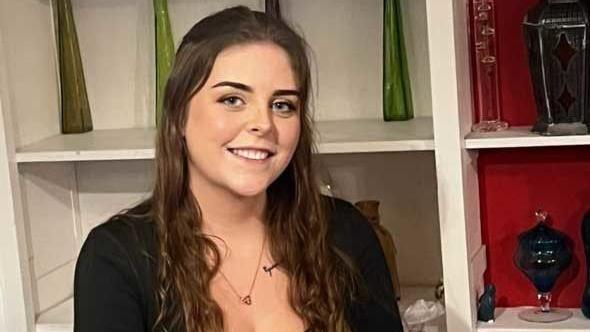 A young woman with long brown hair, wearing a black long sleeved top and a necklace. Behind her are white and red shelves with green and black pottery on them.