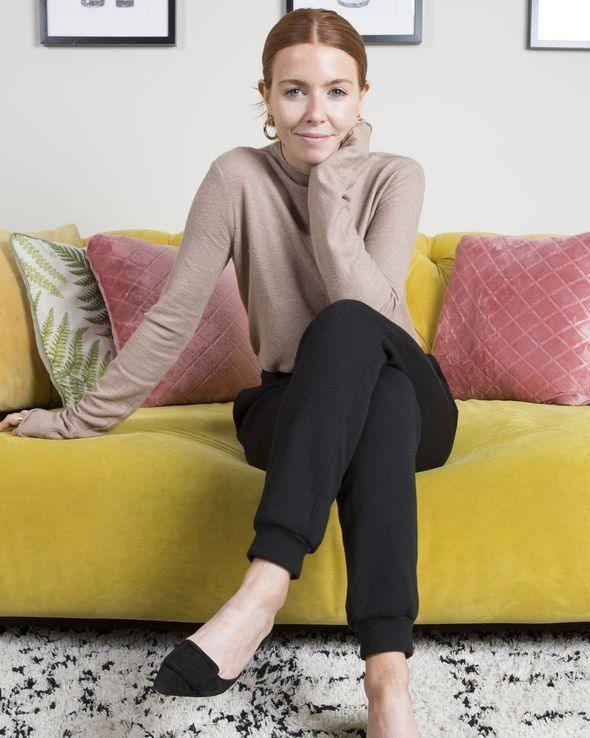 A woman wearing a brown jumper, black trousers and black shoes sits on a bright yellow sofa smiling at the camera. One leg is crossed over the other and her head is resting on her hand.