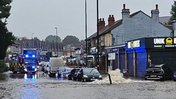 Water is seen gushing from the ground, with water completely flooding a road in the foreground while in the background is a fire engine parked beyond the flood. There is a street with two rows of house and a line of parked cars visible.