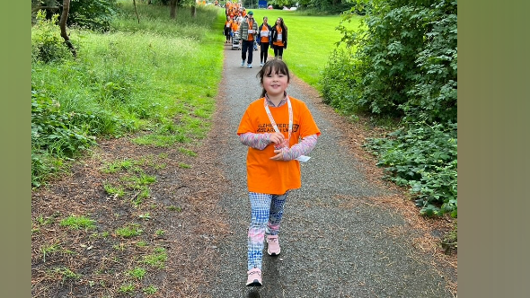 Heidi wears an orange t-shirt with the Alzheimer's Research logo on in black, with blue and pink leggings and pink trainers and is walking through a park on a sponsored walk with people behind her