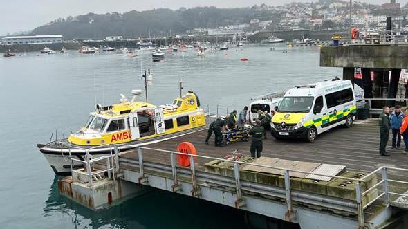 Ambulance crews remove equipment from a yellow marine ambulance on to a pier. Other boats are seen in a harbour, with trees and houses in the background.