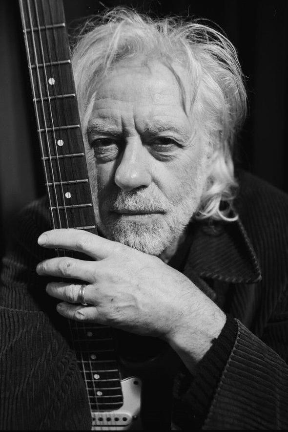 Bob Geldof faces the camera and holds the neck of a guitar with his left hand which has several rings on his ring finger. He has white shaggy layered hair and a white beard