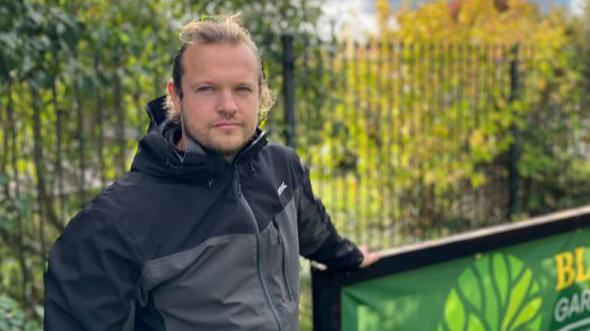 Jordan Holmes has fair tied back hair. He is wearing a black and grey jacket and is leaning against a sign with a fence and shrubs in the background.