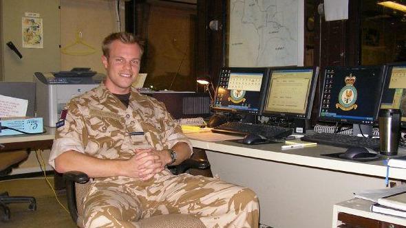 Inside an office at night, Simon King in combat uniform sits next to a desk of computer screen and smiles at the camera