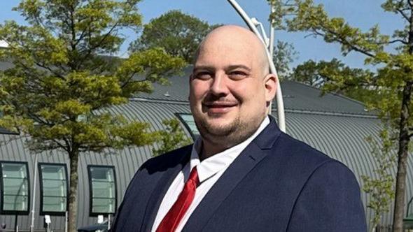 Matt Keane with very short hair and slight beard, wearing a red tie and a blue jacket outside a grey building