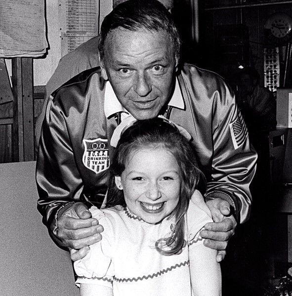 Black and white photo of Frank Sinatra, in a baseball bomber jacket, holding a young Lena Zavaroni's shoulders and leaning over her. Lena is grinning and is wearing a white dress and a big white bow in her hair.