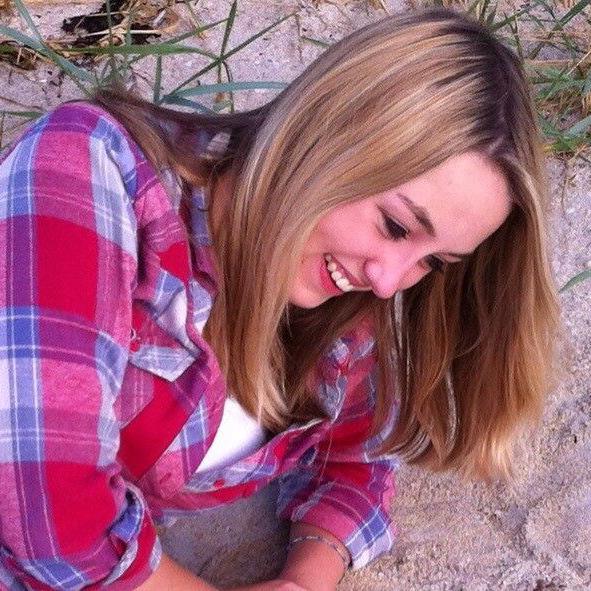 Katie Allan in a happy family photo, looking to be taken at their favourite beach, leaning on the sand with a beaming smile, thick, long blonde hair and wearing a red and blue checked shirt over a white t-shirt. 