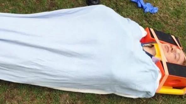 A man lying on a stretcher with a head board to keep his neck straight, covered in a white sheet.