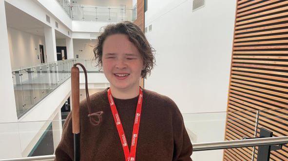 A blind student looking directly at the camera. She is stood in front of a white hall wearing a brown jumper with a red lanyard. She has short hair and in her right hand is her cane that is black with a brown handle at the top. 