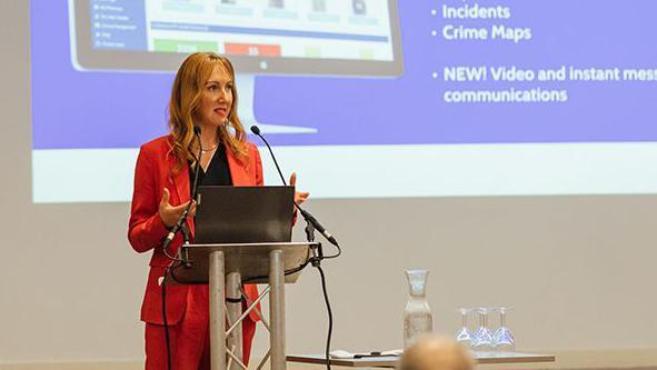 Stephanie Karté dressed in a red blazer and trouser suit delivering a speech as she stands behind a laptop and microphone stand. There is a projector behind her. 