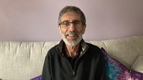 Steve Hobbs - who has a beard and glasses, smiles at the camera while sitting on a sofa.