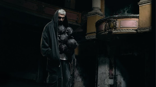 Landscape shot of Throup standing inside the Burnley Empire Theatre. The background is dark and atmospheric and Throup stands wearing a black hoodie and trousers with an avant-garde garment underneath his hoodie