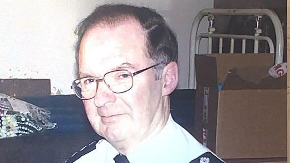 Roger Cook, wearing glasses and a black and white uniform, looks toward the camera